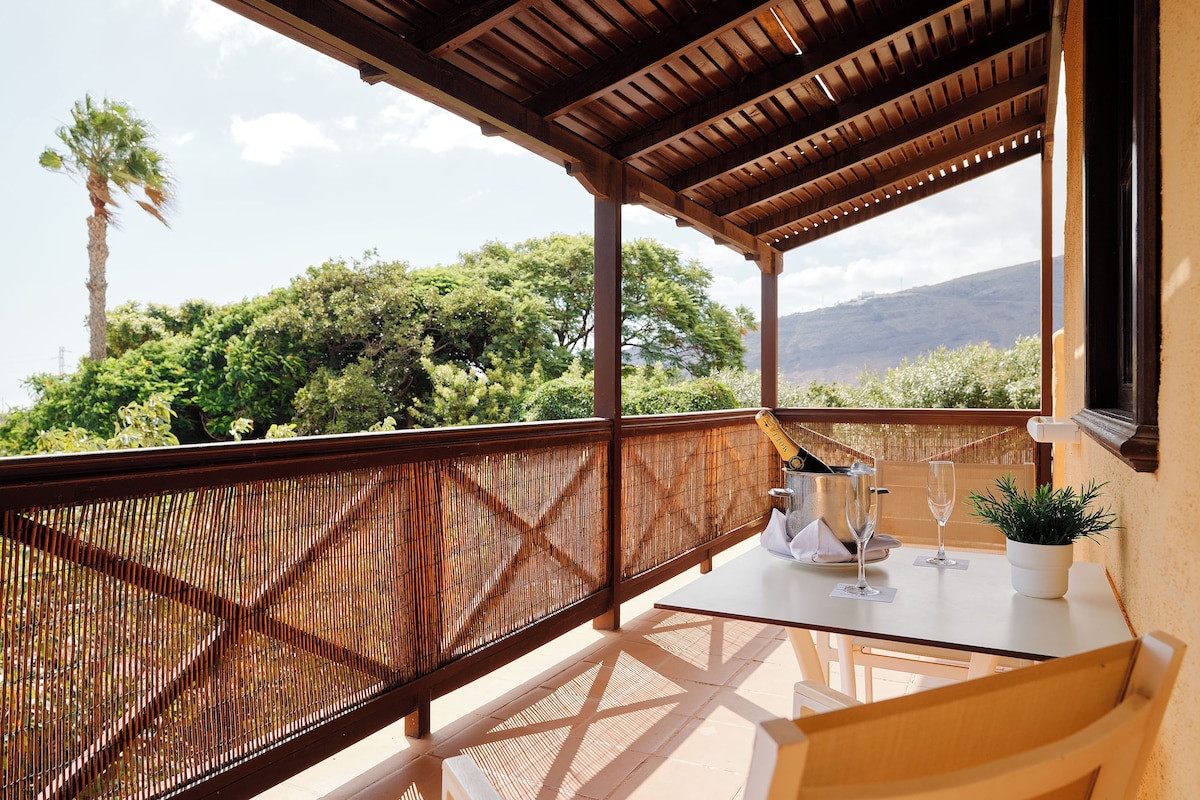 A covered outdoor area with a wooden railing, a table with a bottle and glasses, and a view of greenery and hills.