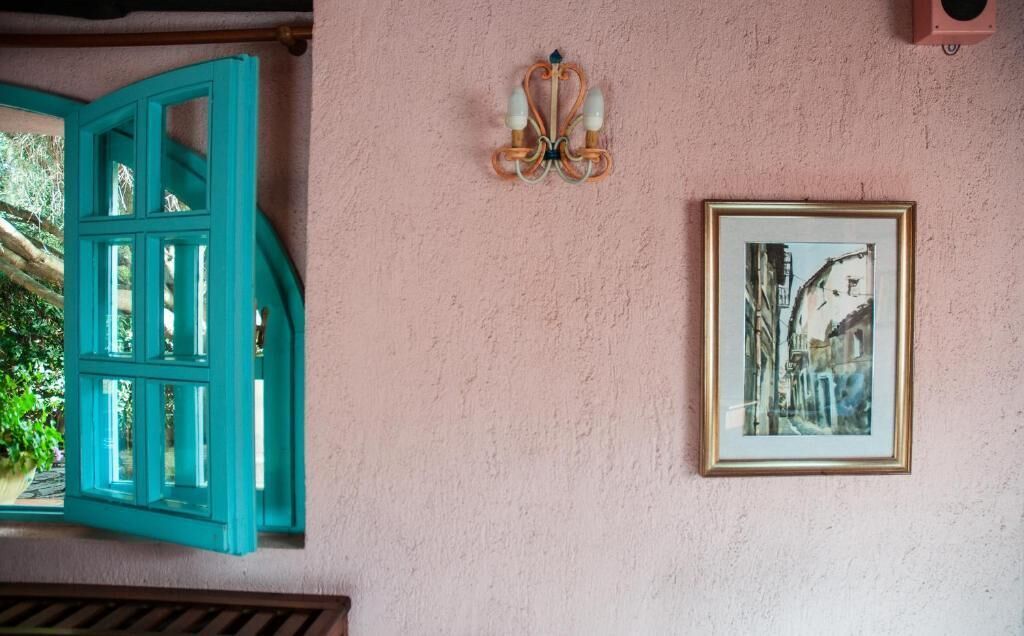 Een roze muur met een ingelijst schilderij, een raam met een blauwgroene lijst en een wandlamp.