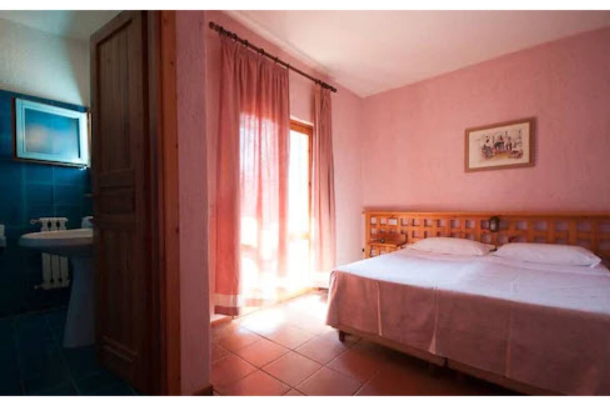 A bedroom with a bed, a nightstand, a window with curtains, a bathroom with a sink and mirror, and a framed picture on the wall.