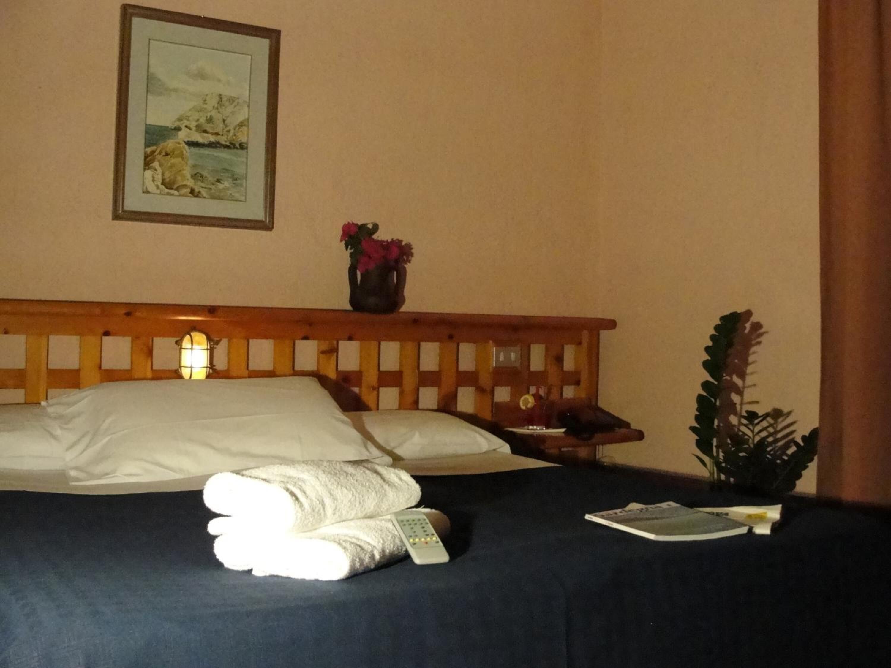 A neatly made bed with a white pillow, a folded towel, and a remote control. There is a small table with a vase of flowers and a glass of water. A framed picture hangs on the wall above the bed.