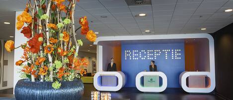 Un área de recepción con un jarrón azul grande lleno de flores anaranjadas, un mostrador de recepción y una pantalla digital que muestra la palabra 'RECEPTION'.