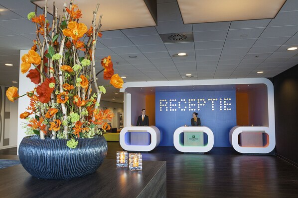 Un área de recepción con un jarrón azul grande lleno de flores anaranjadas, un mostrador de recepción y una pantalla digital que muestra la palabra 'RECEPTION'.