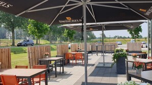 An outdoor patio area with tables and chairs under umbrellas, a view of the road, and a fence. - Fletcher Wellness - Hotel Sittard (Sittard)