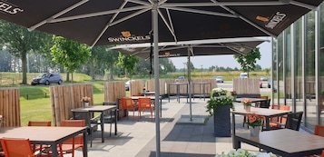 Un patio exterior con mesas y sillas bajo sombrillas, vista a la carretera y una cerca.