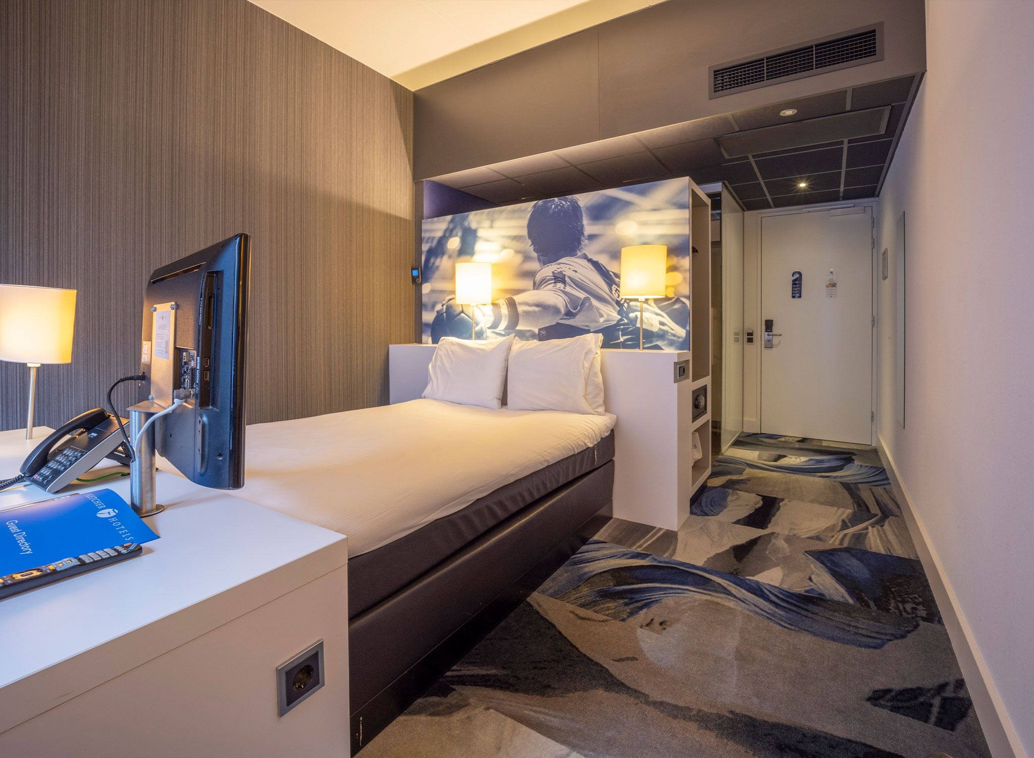 A hotel room with a bed, a desk with a phone and a directory, a wall-mounted lamp, and a door leading to a bathroom.
