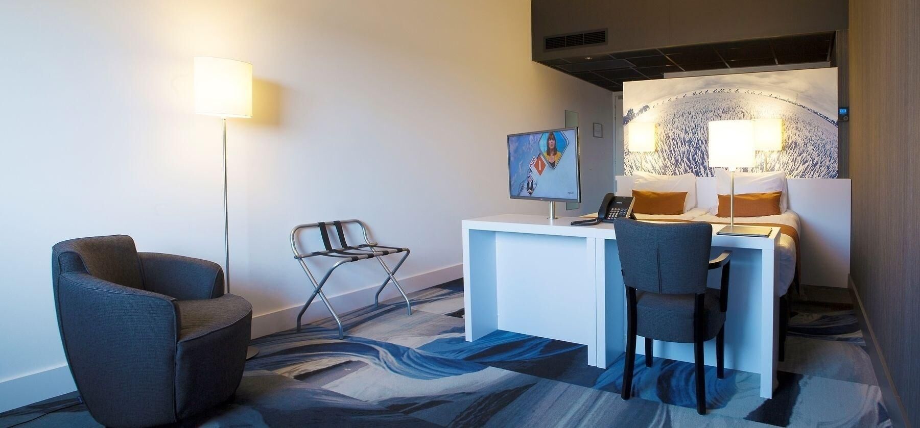 A hotel room with a desk, two chairs, a bed with pillows, a wall-mounted TV, and a large abstract painting.