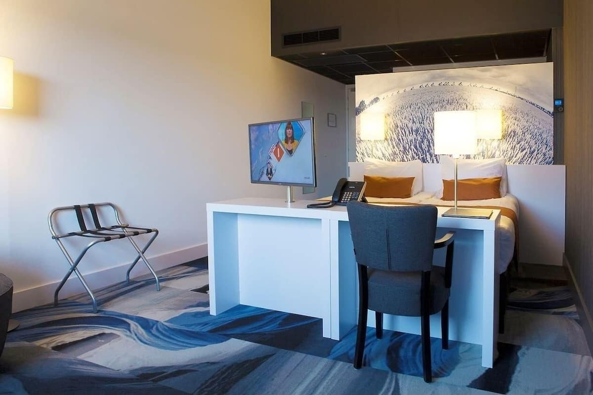 A hotel room with a desk, two chairs, a bed with pillows, a wall-mounted TV, and a large abstract painting.