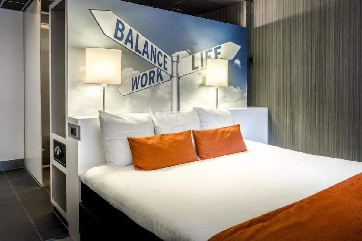 A modern hotel room with a bed, pillows, and a wall art piece depicting 'BALANCE LIFE WORK'.