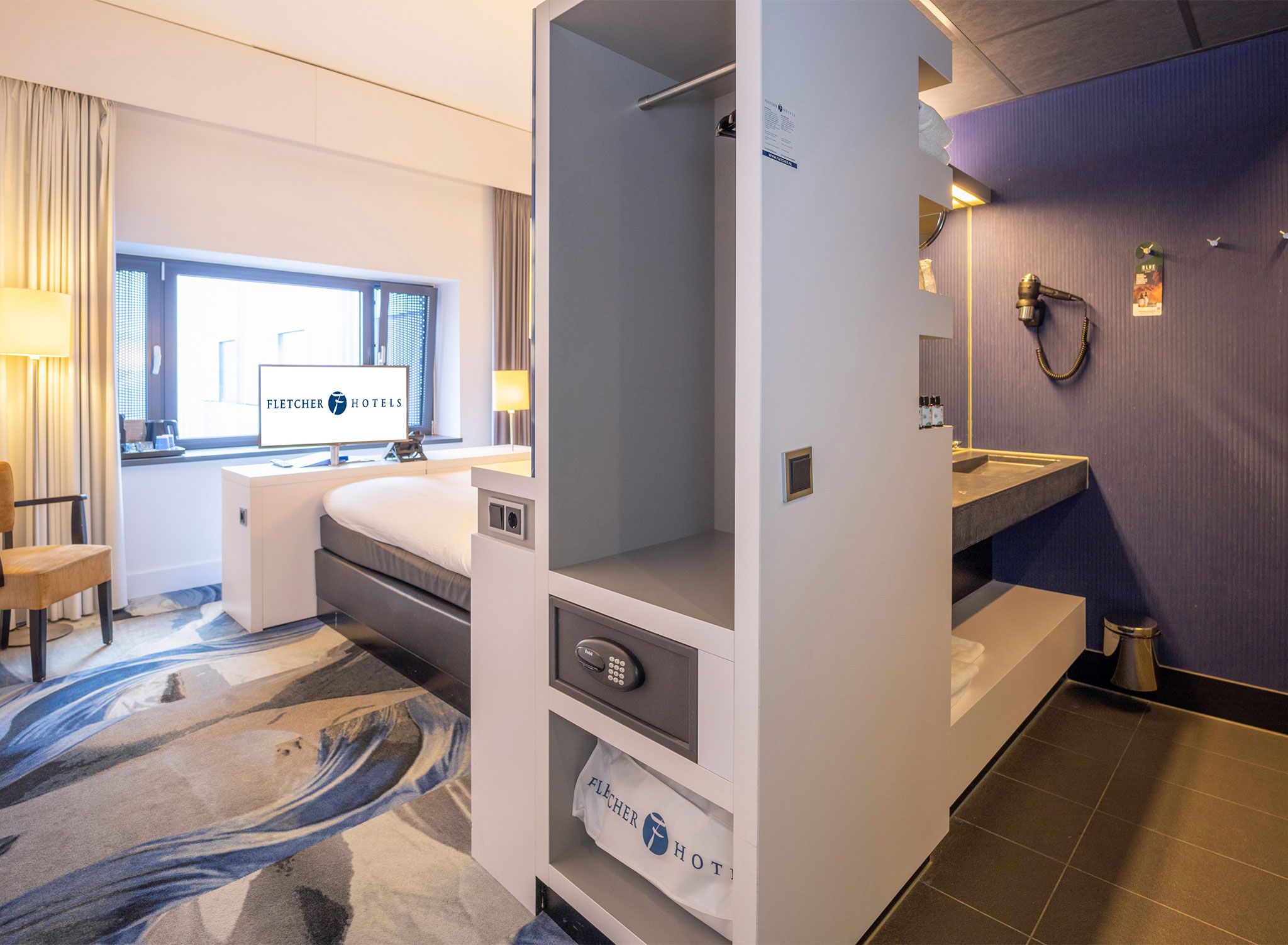 Una camera d'albergo con un letto, una scrivania, un bagno con lavandino e specchio, e una finestra con tende.