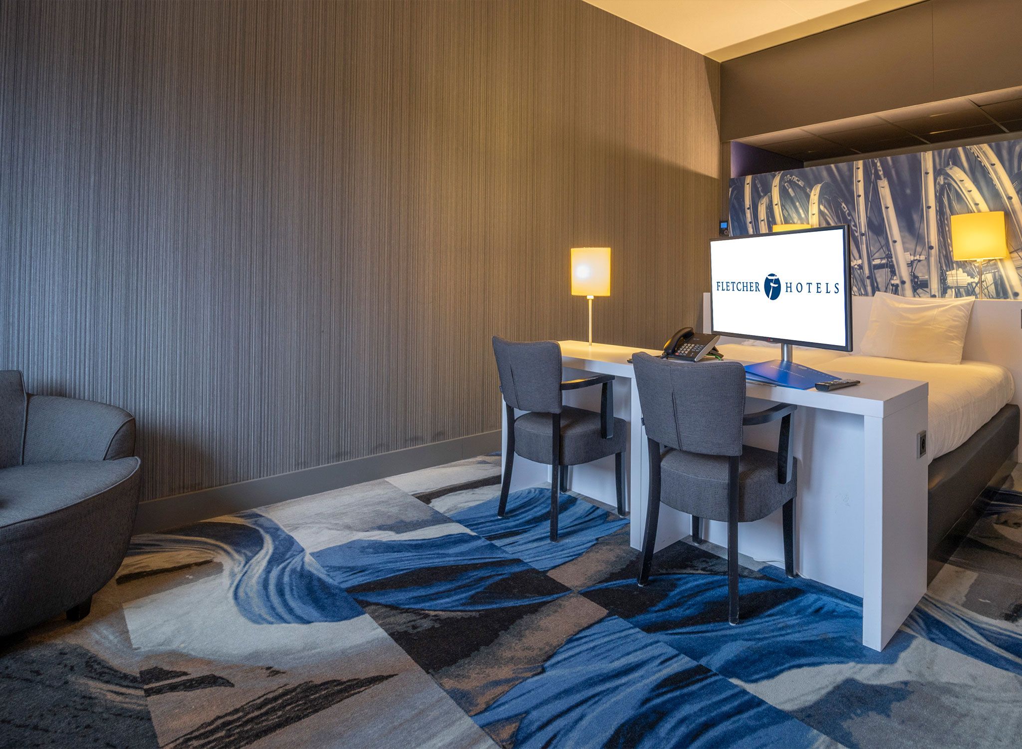 A hotel room with a bed, a desk with two chairs, a computer, and a wall-mounted TV displaying the hotel's name.