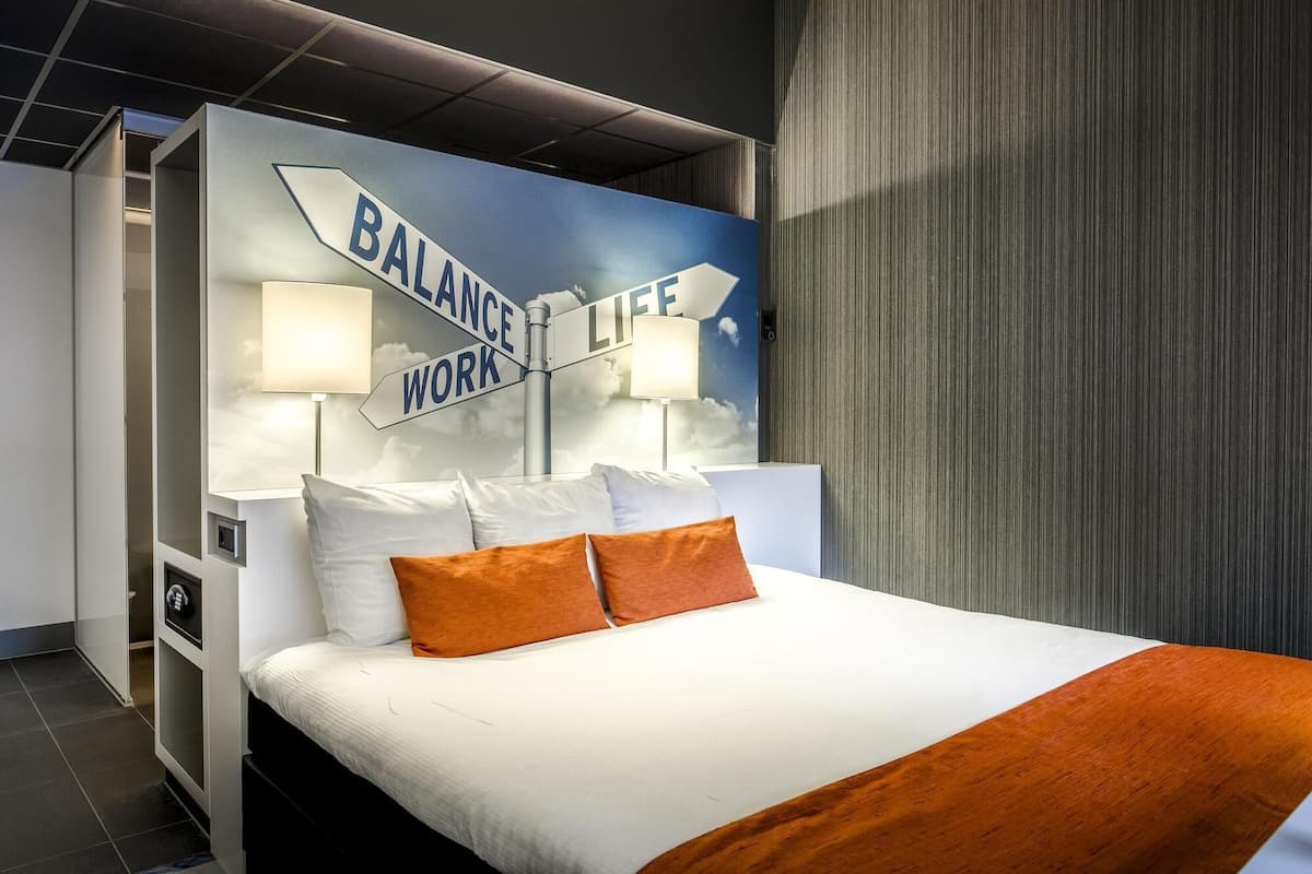 Ett hotellrum med en stor säng, vita sängkläder och två orange kuddar. På väggen sitter en skylt med texten "BALANSERA LIV OCH ARBETE".