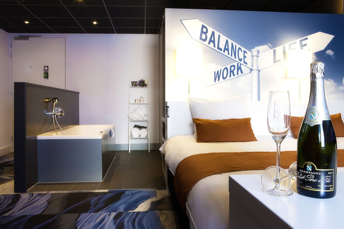 A hotel room with a bed, a nightstand with a bottle of champagne and a glass, a bathroom with a bathtub, and a sign that reads "Balance Life Work".