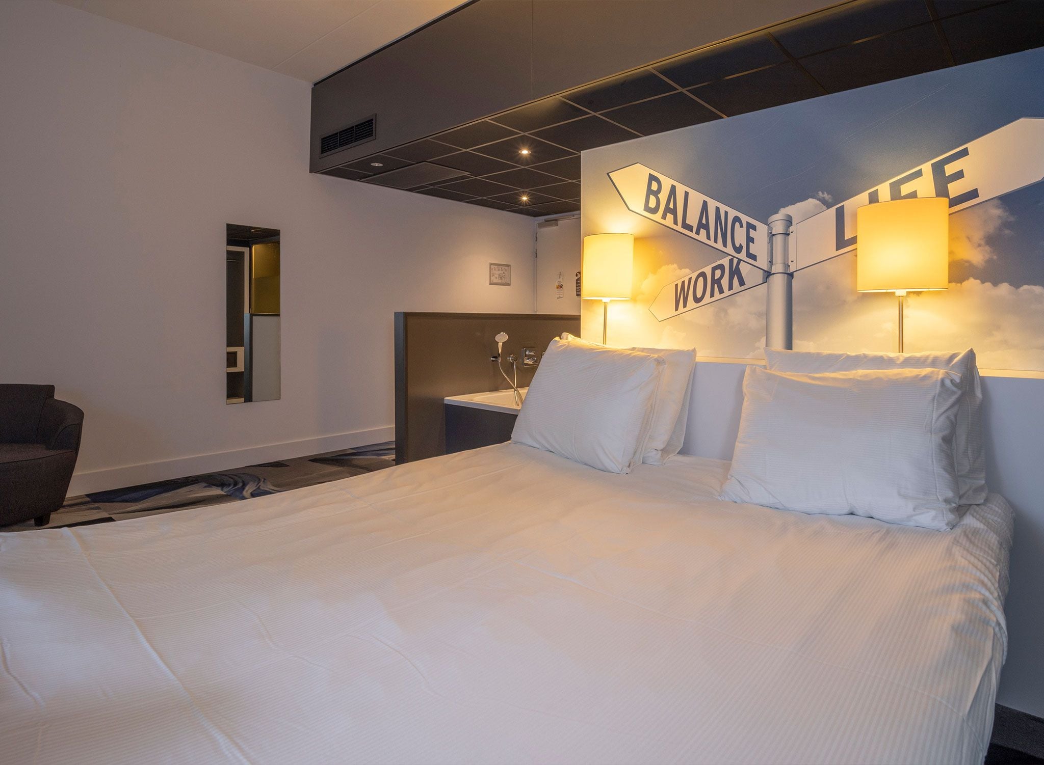 A hotel room with a large bed, a headboard with 'BALANCE WORK' signage, a nightstand, a chair, and a shower area.