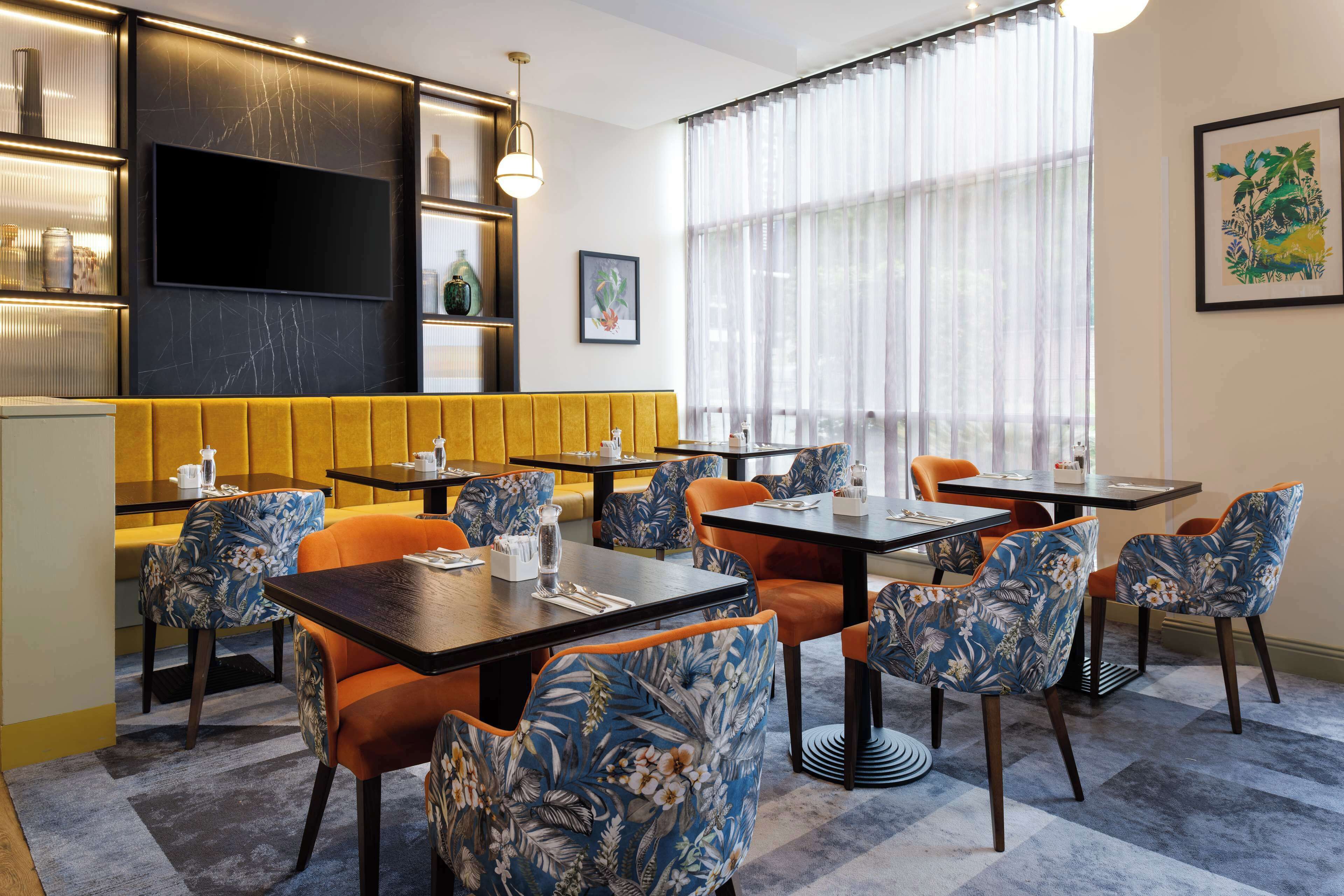 A restaurant with a yellow booth seating area, tables with blue floral chairs, and a large window with sheer curtains.