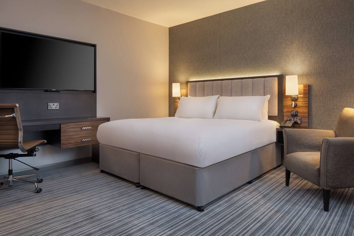A modern hotel room with a large bed, a flat-screen TV, a desk with a chair, and two bedside lamps.