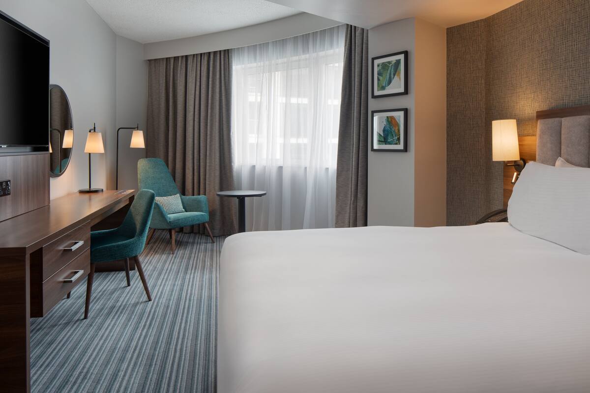 A hotel room with a large bed, a desk with a chair, a TV, two lamps, and a window with curtains.
