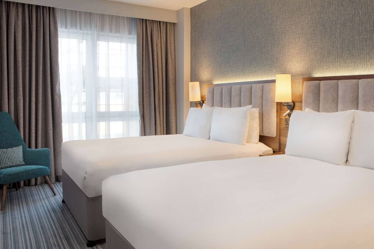 A hotel room with two beds, a teal armchair, and a large window with curtains.