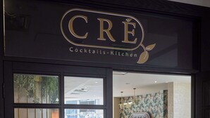 Entrada a un restaurante con un letrero que dice "CRE Cocktails-Kitchen". En el interior, hay varias mesas y sillas dispuestas para comer.