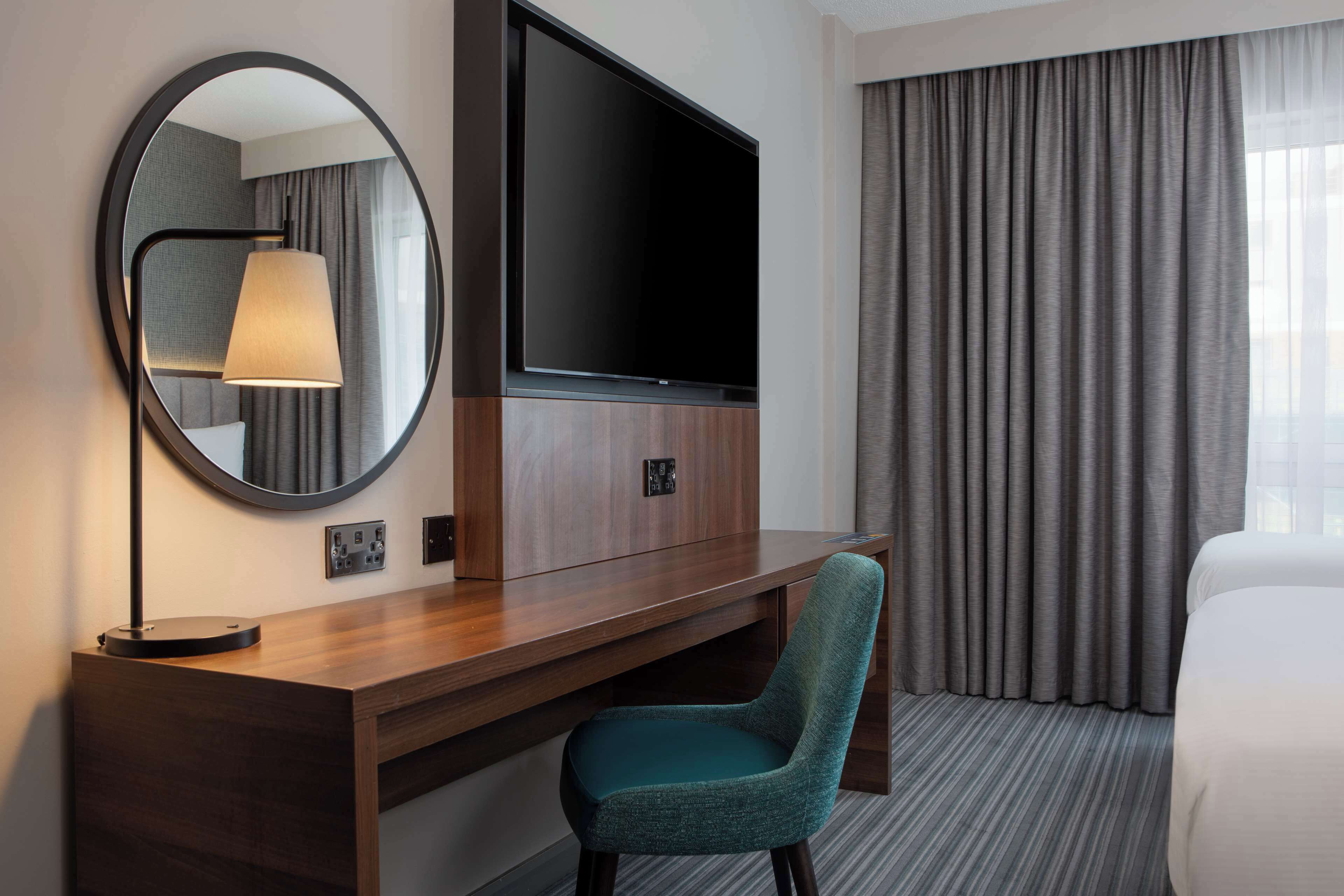 A hotel room with a wooden desk, a flat-screen TV, a round mirror, a teal chair, a large window with curtains, and a bed.