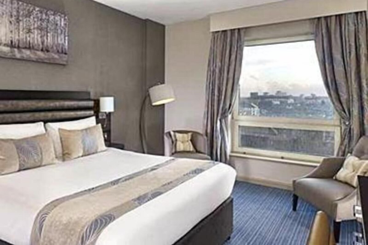 A hotel room with a large bed, a chair, a lamp, and a view of the city.