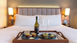 Habitación de hotel con una cama bien hecha, una bandeja con comida y vino, y una mesita de noche con una lámpara y teléfono.