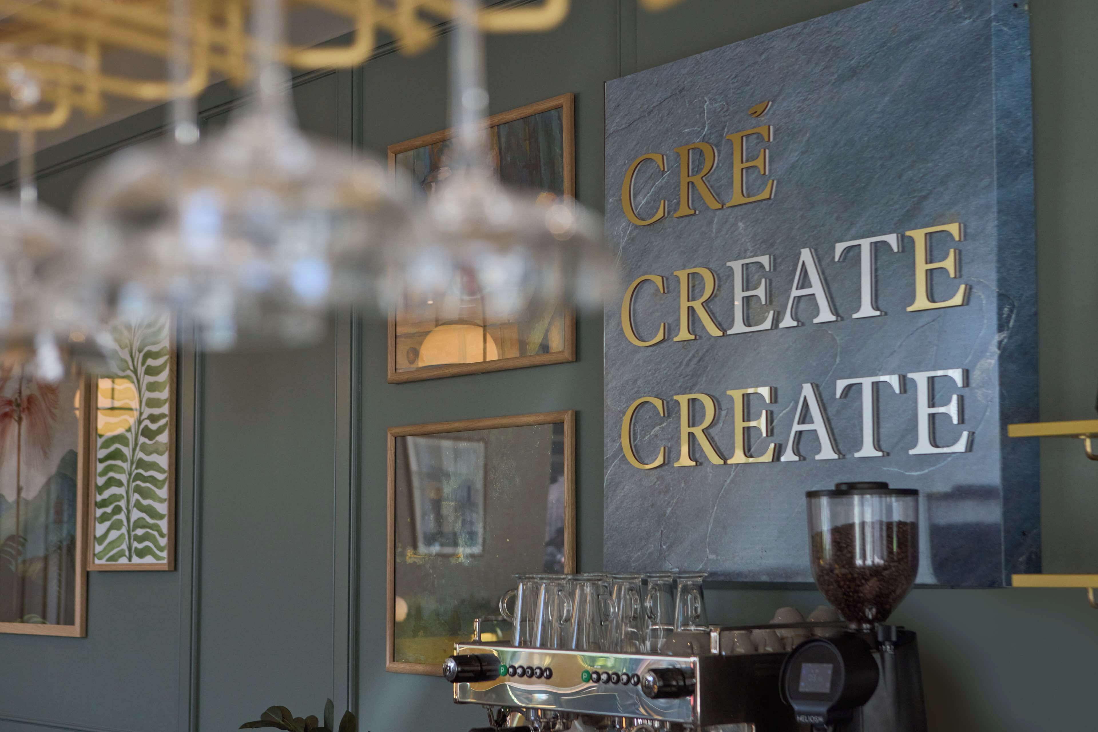 A coffee machine with a grinder and a glass carafe, a wall with framed artwork, and a sign that reads "CRÉ CREATE CREATE".