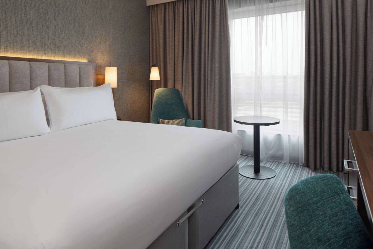A hotel room with a large bed, a desk, a chair, a small table, and a lamp.