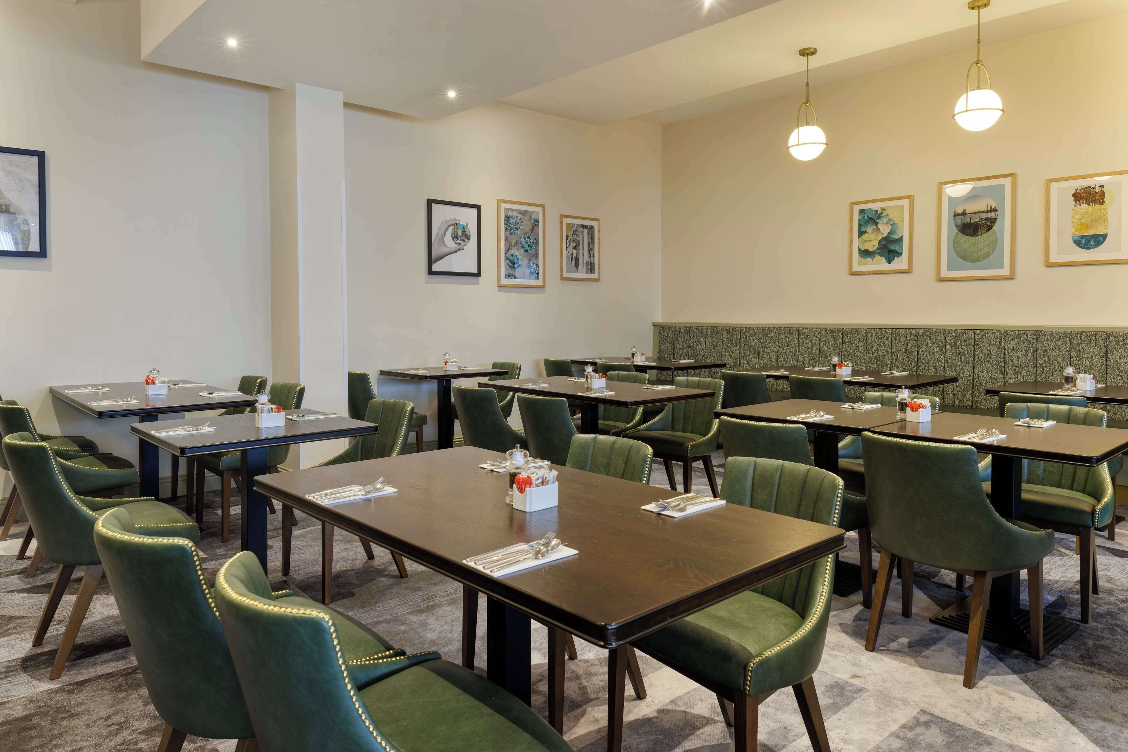 A hotel conference room with multiple tables and green upholstered chairs, framed artwork on the walls, and a carpeted floor.