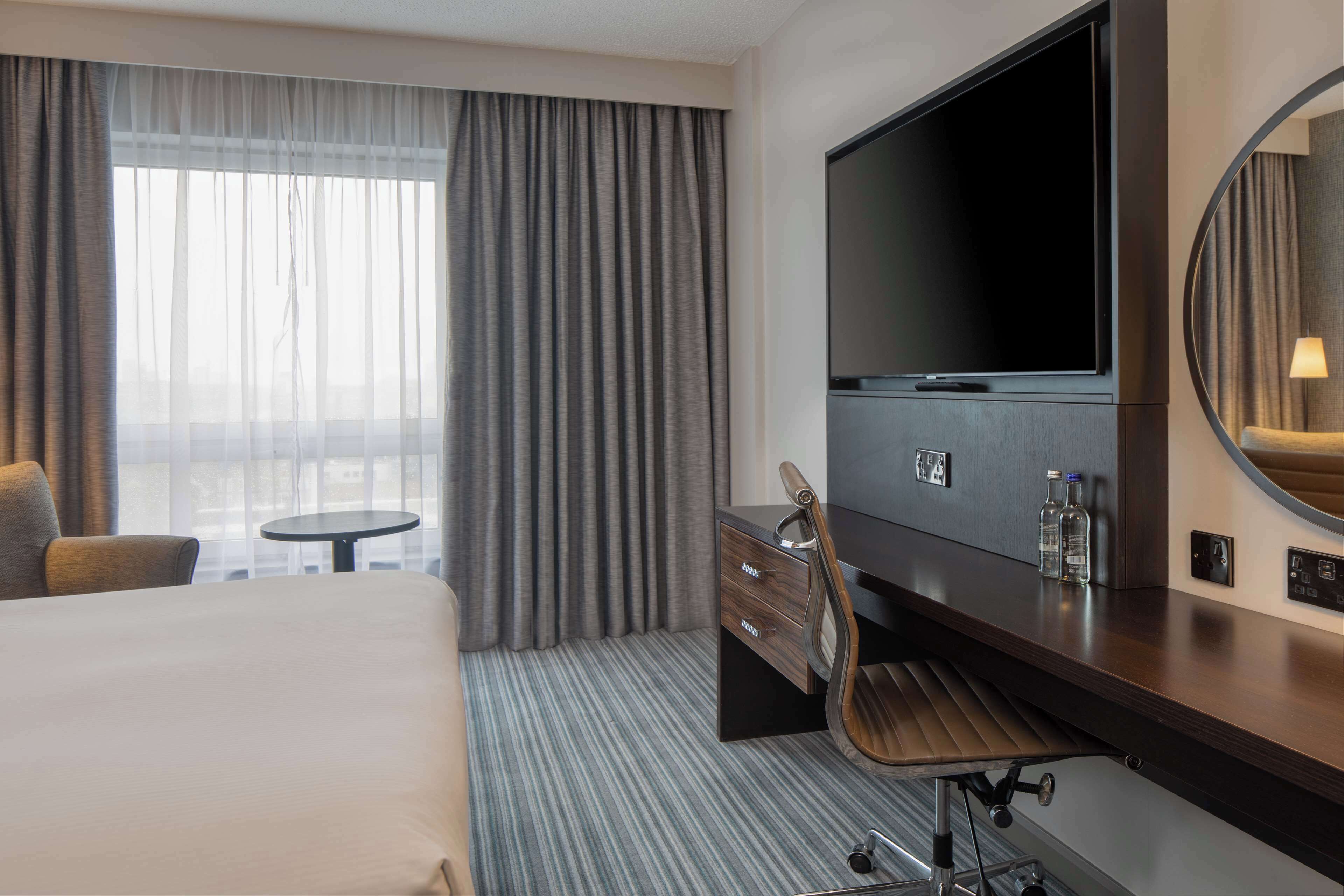 Ein Hotelzimmer mit einem großen Bett, einem Schreibtisch mit Flachbildfernseher, einem Sessel, einem kleinen runden Tisch und einem großen Fenster mit Vorhängen.
