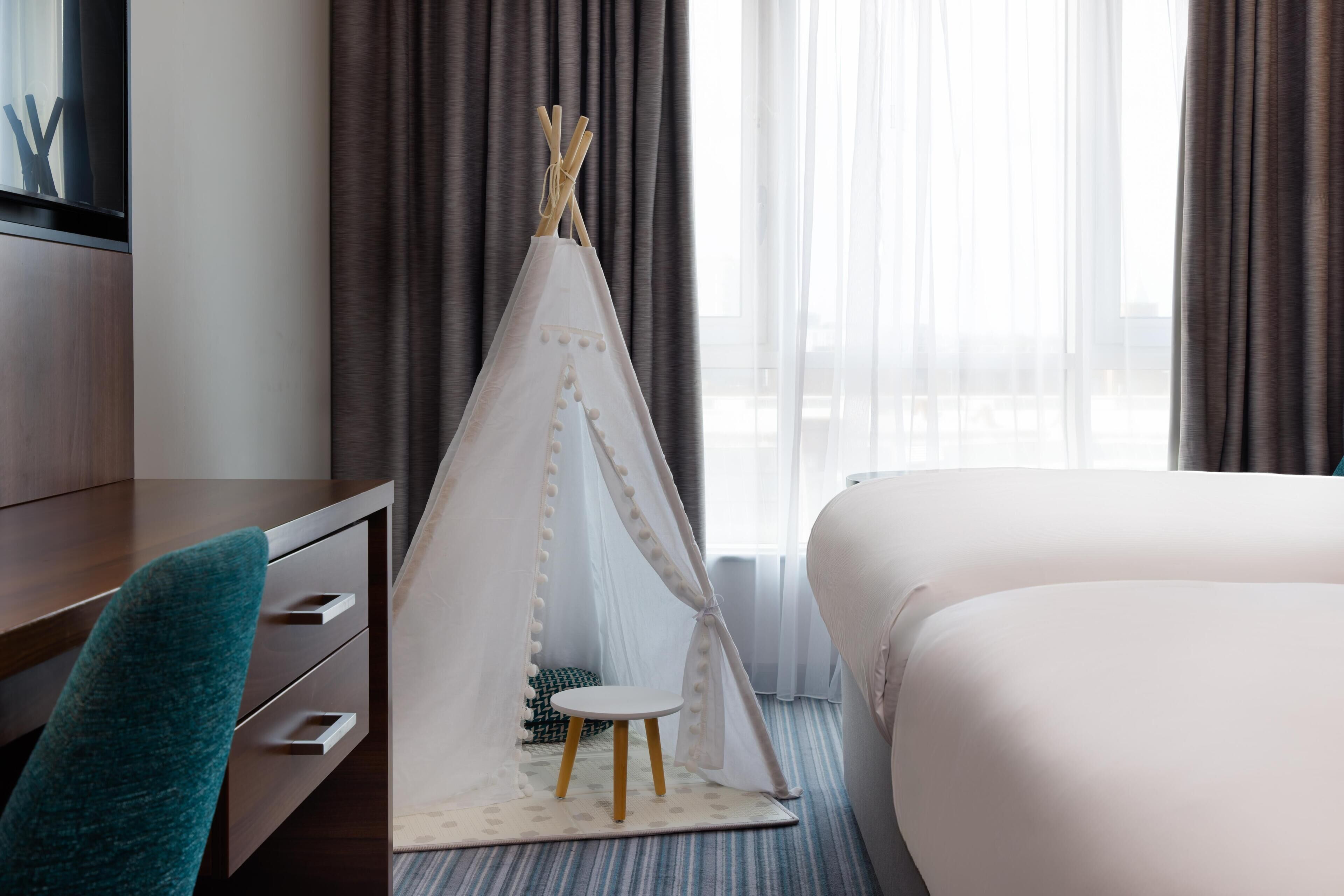 A hotel room with a bed, a desk with drawers, a chair, a small round table, and a teepee-like structure with sticks.