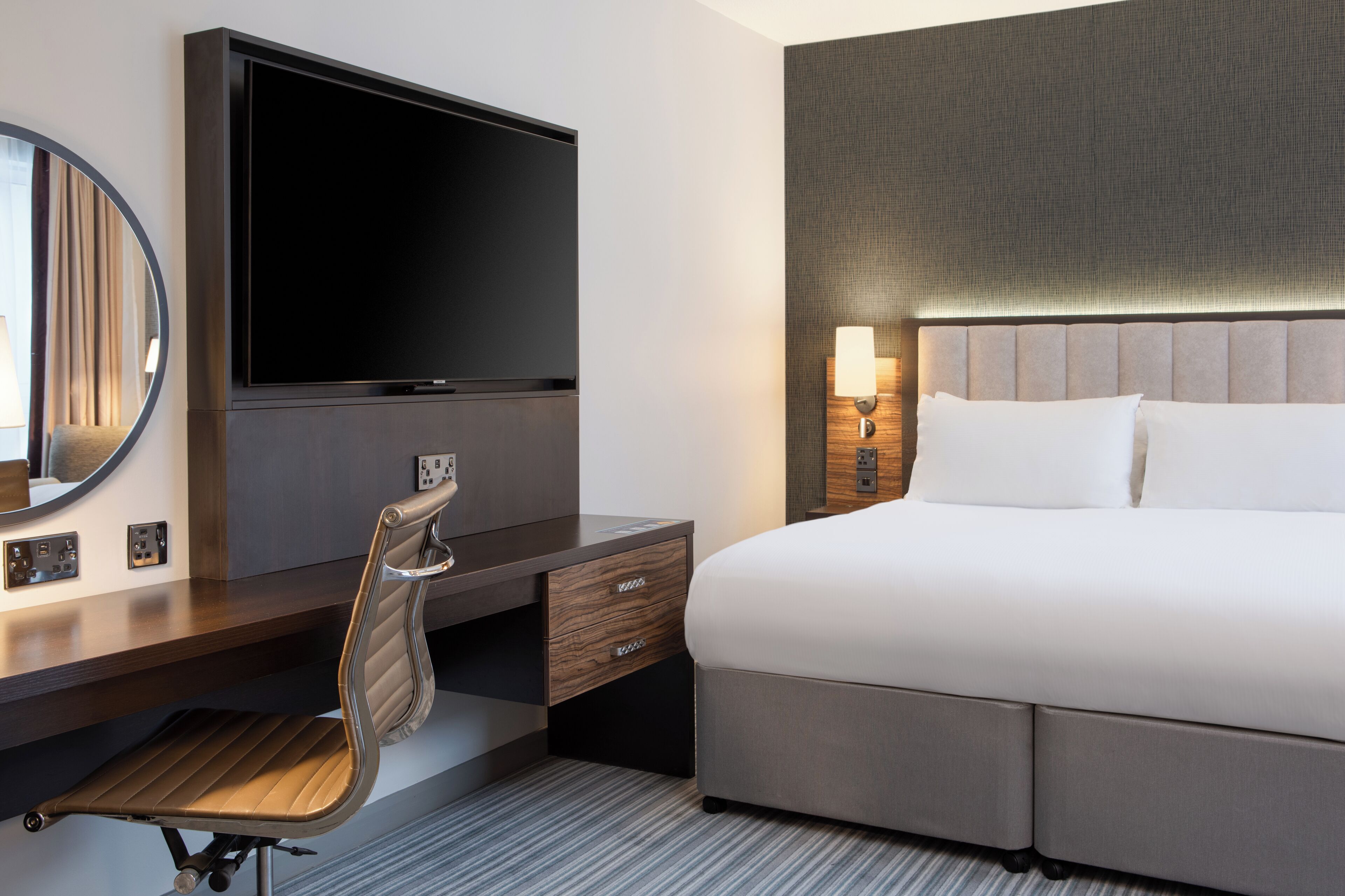 A hotel room with a large bed, a flat-screen TV mounted on a dark wooden dresser, a desk with a chair, and a wall-mounted lamp.