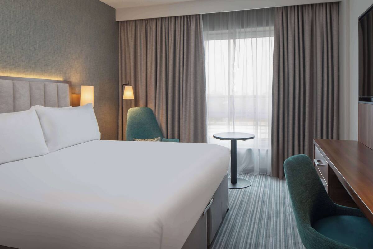 A hotel room with a large bed, a desk, a chair, a small table, and a view through the window.