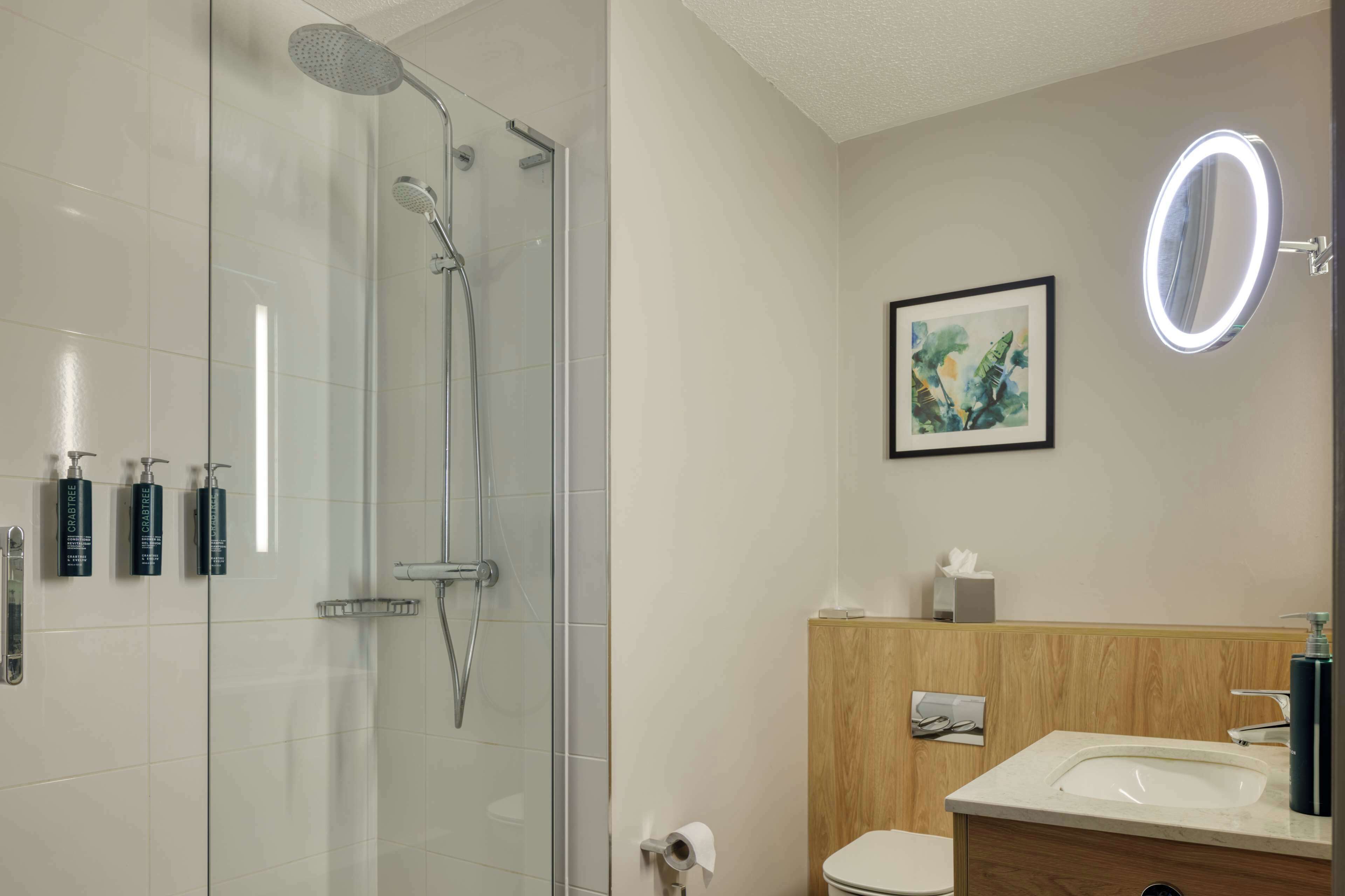Un baño moderno con cabina de ducha de vidrio, tocador de madera, lavabo y una obra de arte enmarcada en la pared.