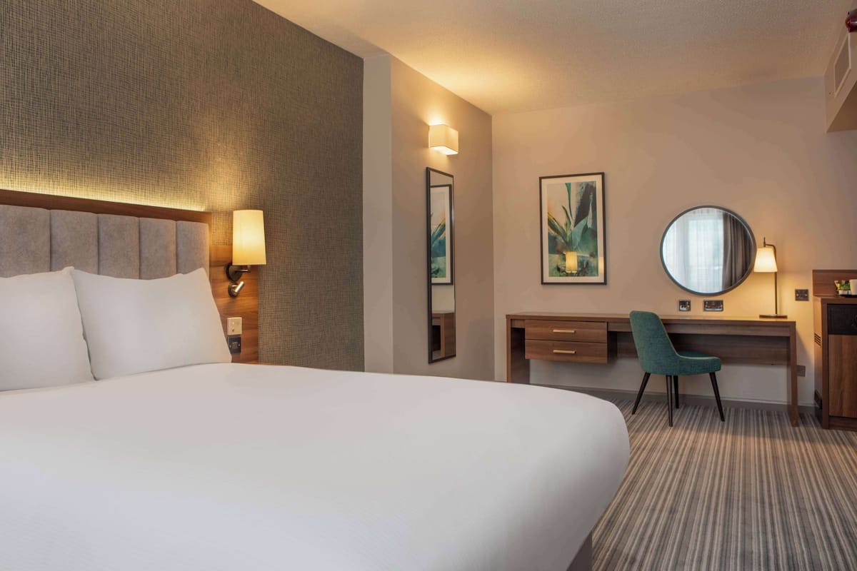 A hotel room with a large bed, a desk with a chair, a round mirror, and two wall-mounted lamps.