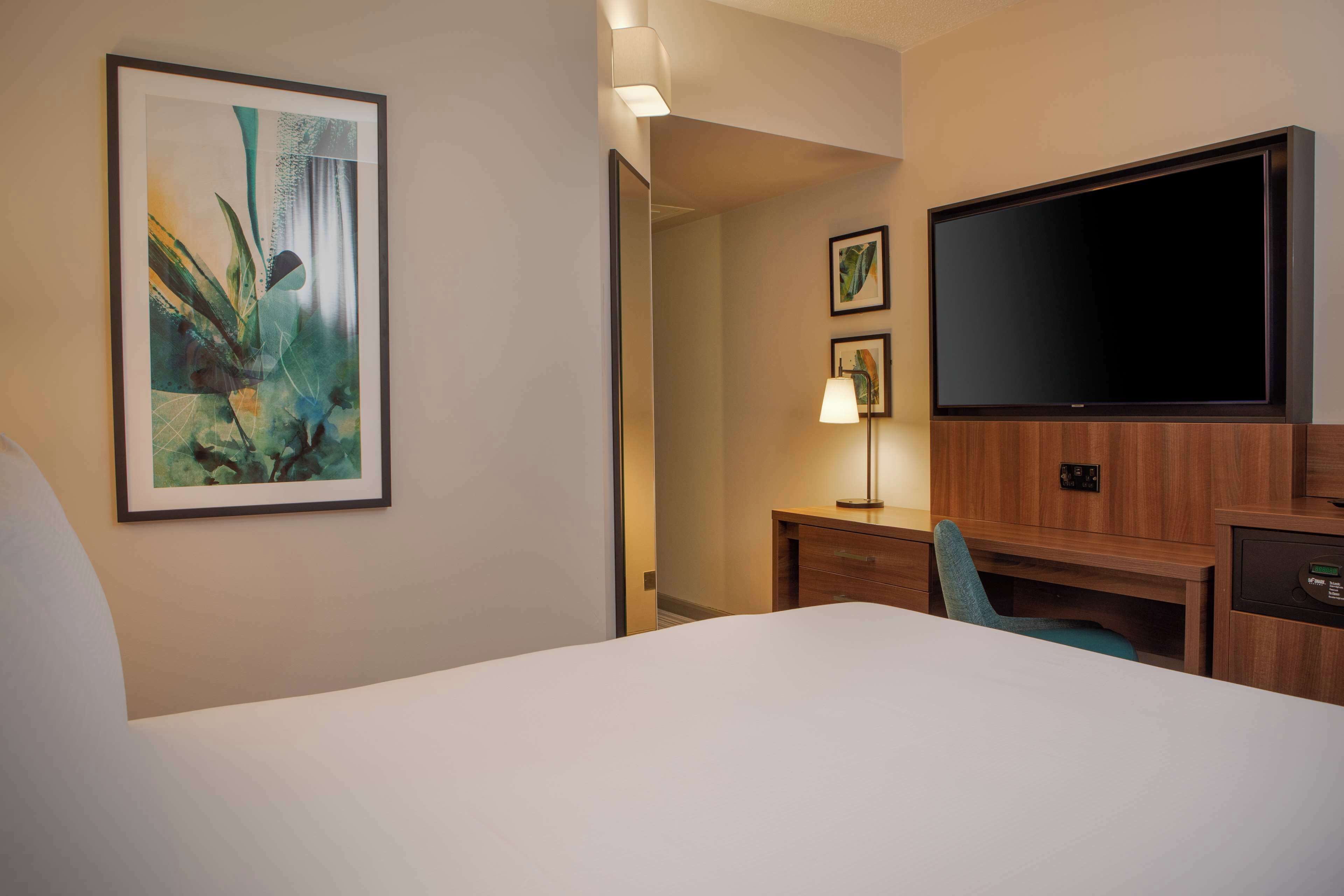 A hotel room with a bed, a desk, a TV, and a painting on the wall.