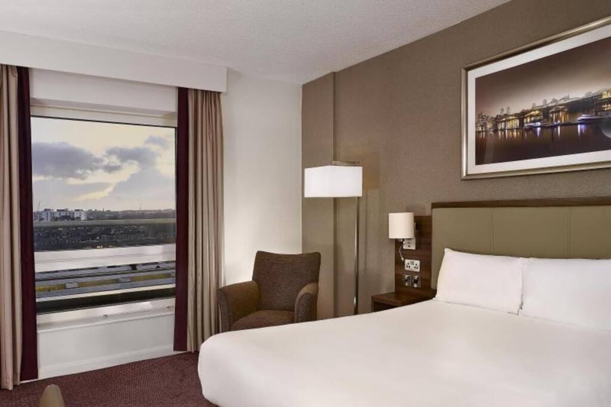 A hotel room with a large bed, a chair, a nightstand with a lamp, a window with curtains, and a cityscape view.
