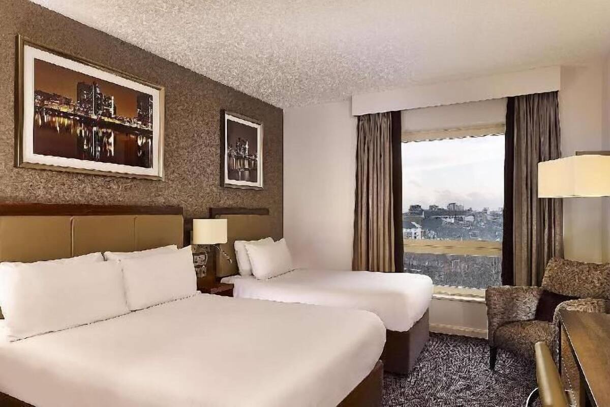A hotel room with two beds, a desk, a chair, a television, and a large window with a city view.