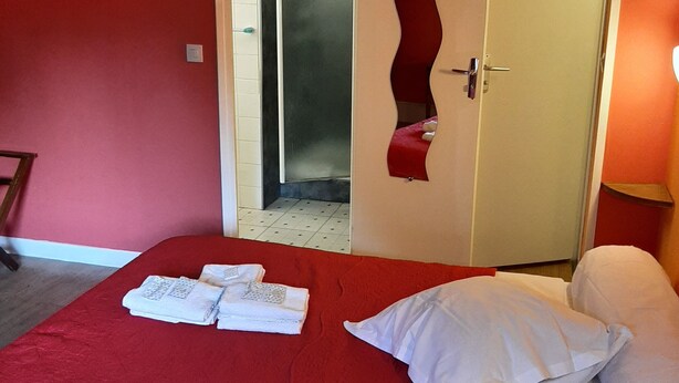 A hotel room with a red bedspread, white pillows, a neatly made bed with towels, a door with a unique design, and a visible bathroom. - Hotel du Cygne (Beauvais)