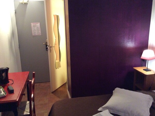 Quarto de hotel com uma cama, uma escrivaninha com cadeira, uma mesa com cadeira, um abajur e uma porta.