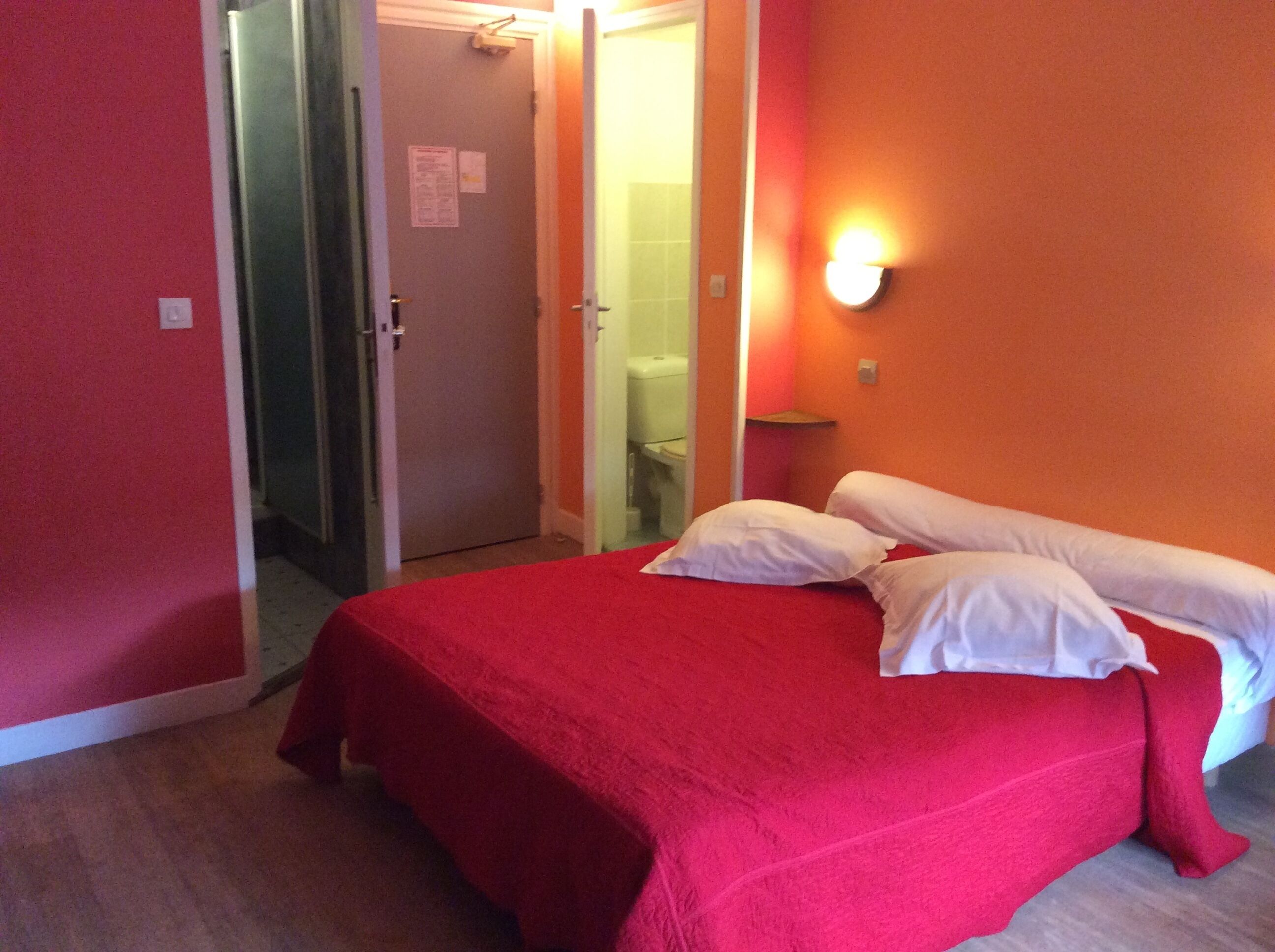 Una habitación de hotel con cama, pared roja y baño visible a través de una puerta abierta.