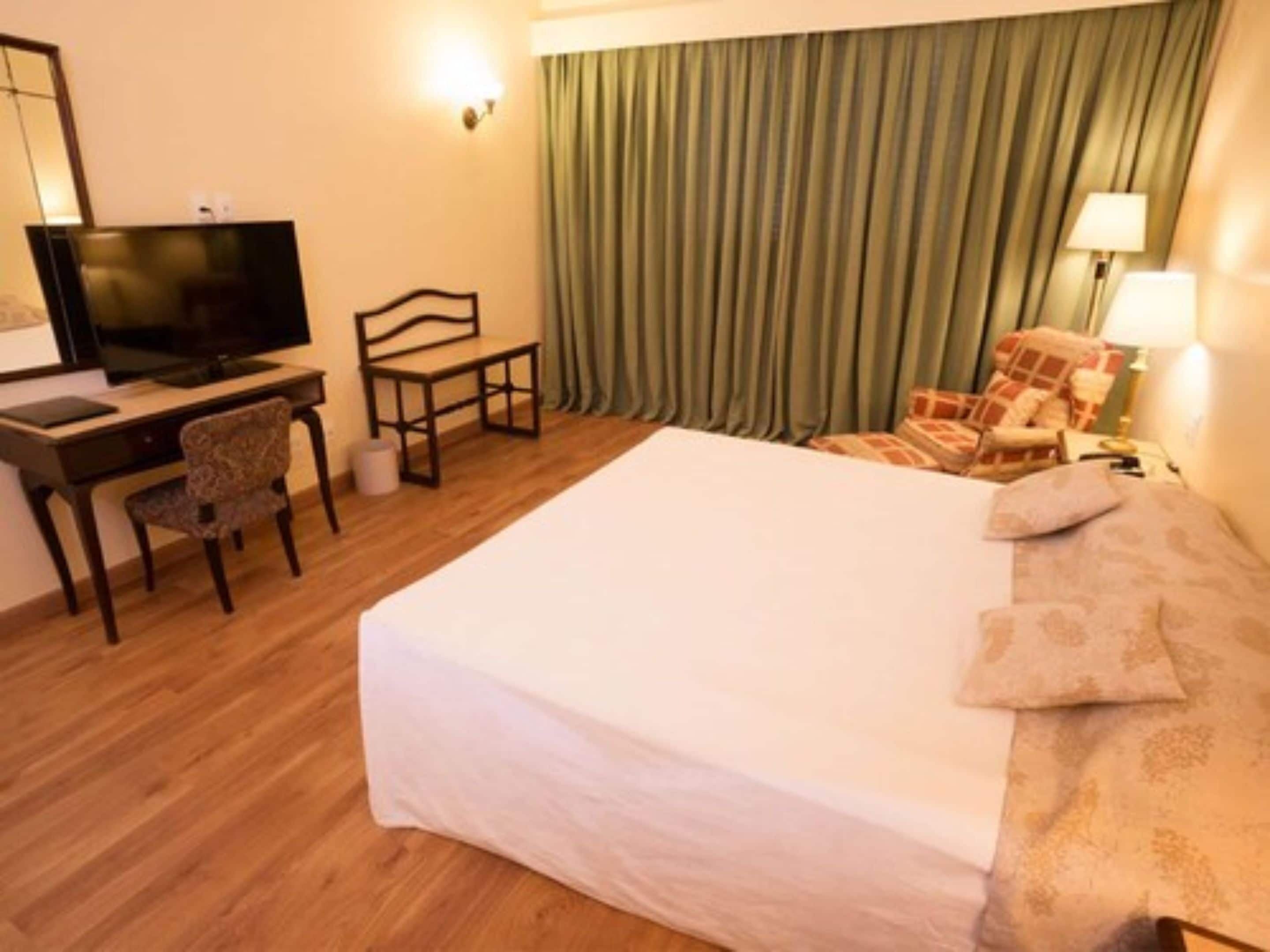 Une chambre d’hôtel avec un lit, un bureau avec une télévision, une chaise, une lampe et un rideau.