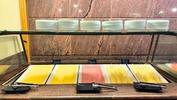 Buffet com diversas opções de comida, incluindo pratos amarelos e cor-de-rosa, e uma vitrine de vidro com pratos empilhados.