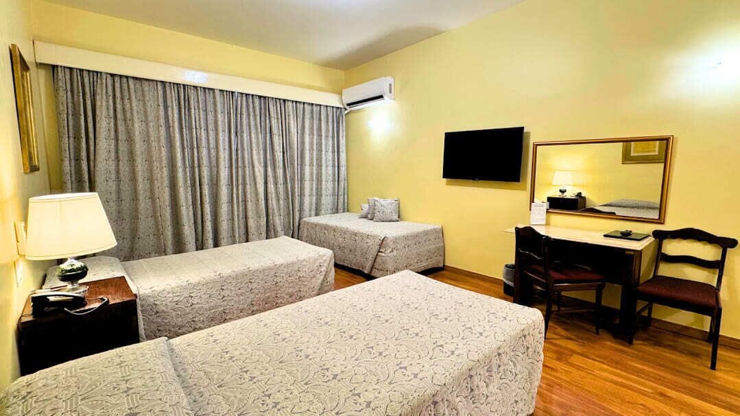 A hotel room with two beds, a desk, a chair, a TV, and an air conditioning unit.
