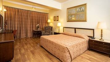 Quarto de hotel com uma cama grande, piso de madeira, escrivaninha, uma cadeira e uma mesa com abajur.