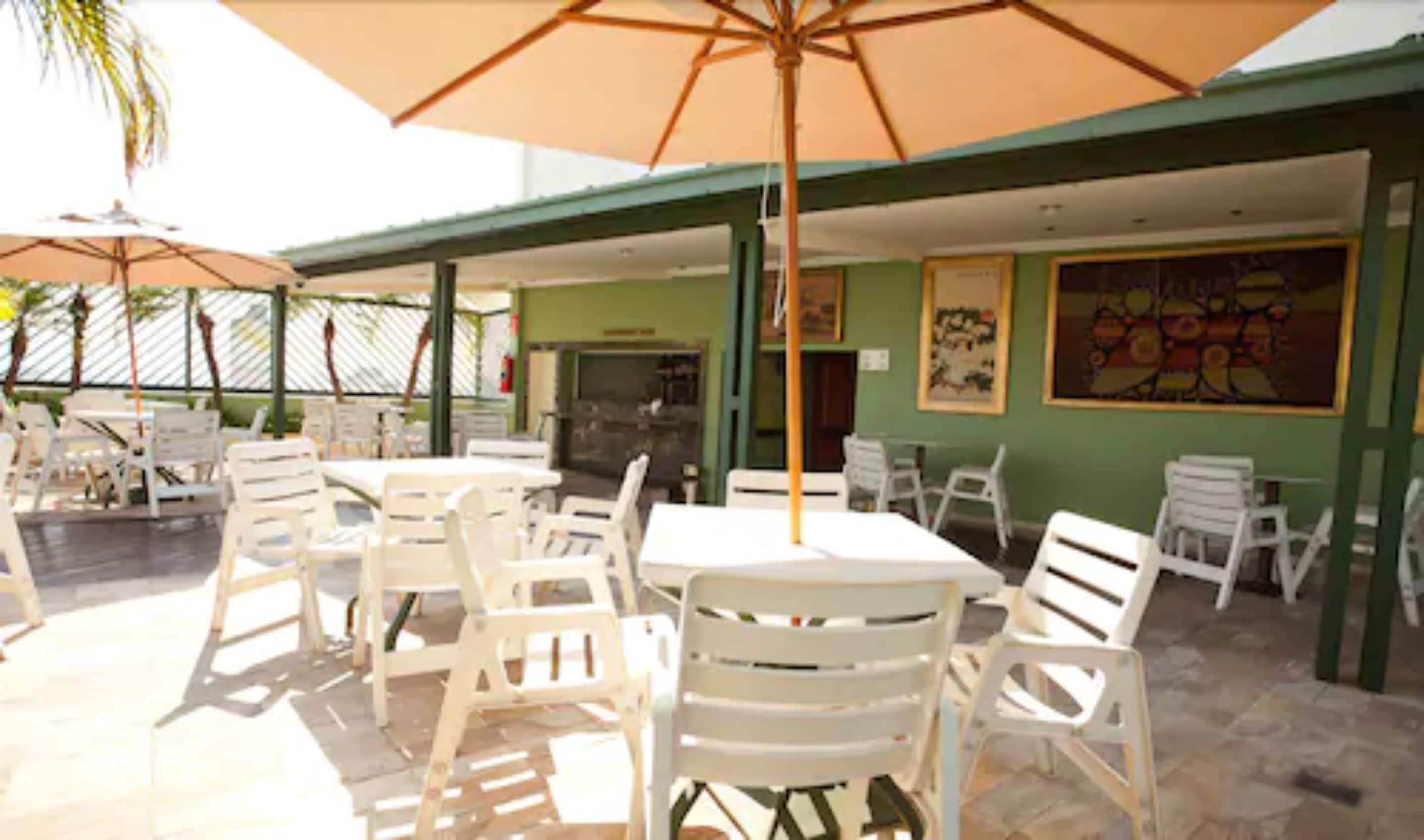 Un espace extérieur aménagé en terrasse, avec des chaises et des tables en plastique blanc, protégé par de grands parasols, et un mur végétal agrémenté d’œuvres d’art.