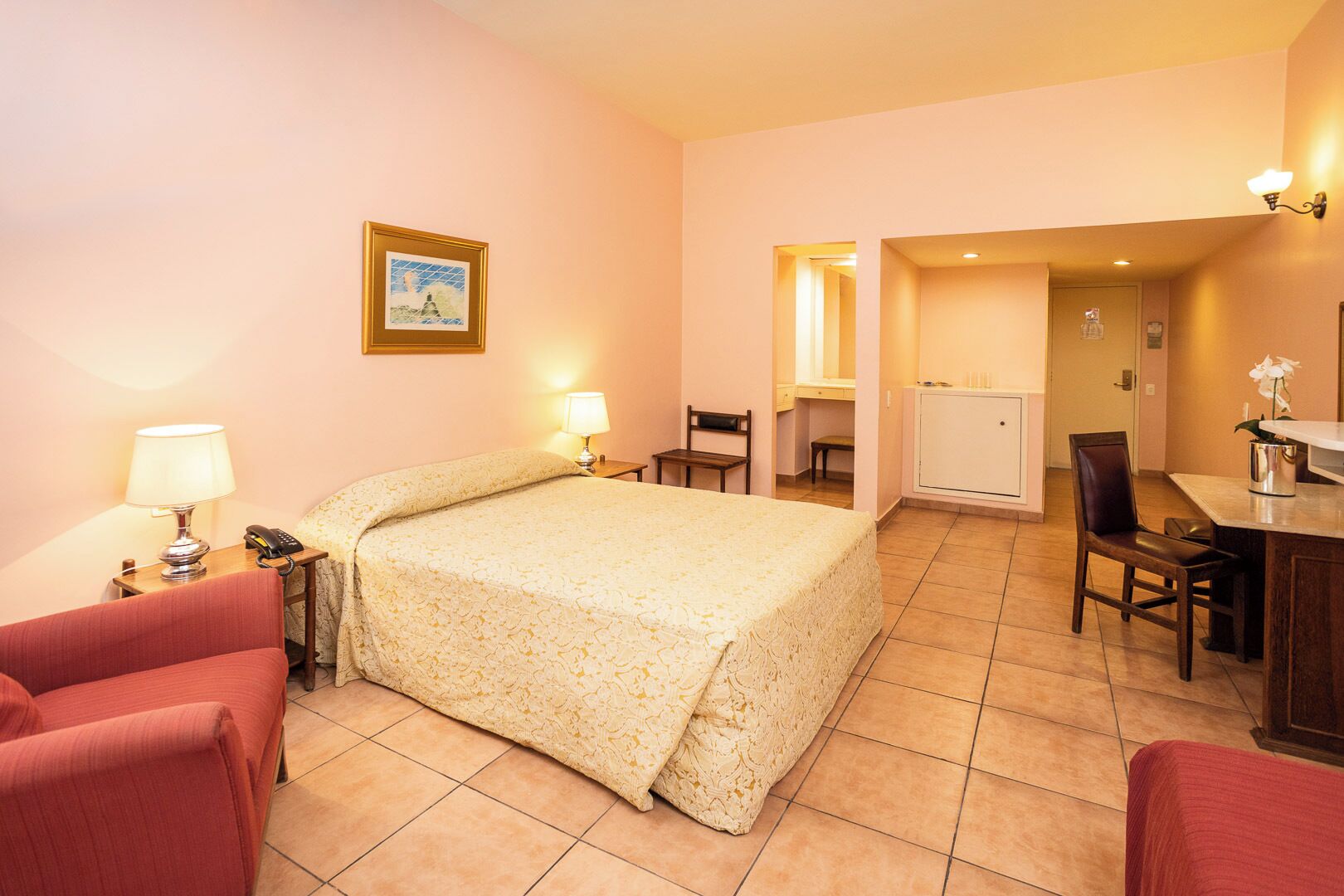 A hotel room with a bed, two chairs, a desk, a lamp, a phone, a picture on the wall, and a small cabinet.