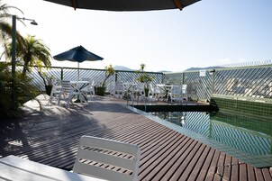 Un área junto a la piscina con sillas reclinables blancas, una terraza de madera y vista al mar.
