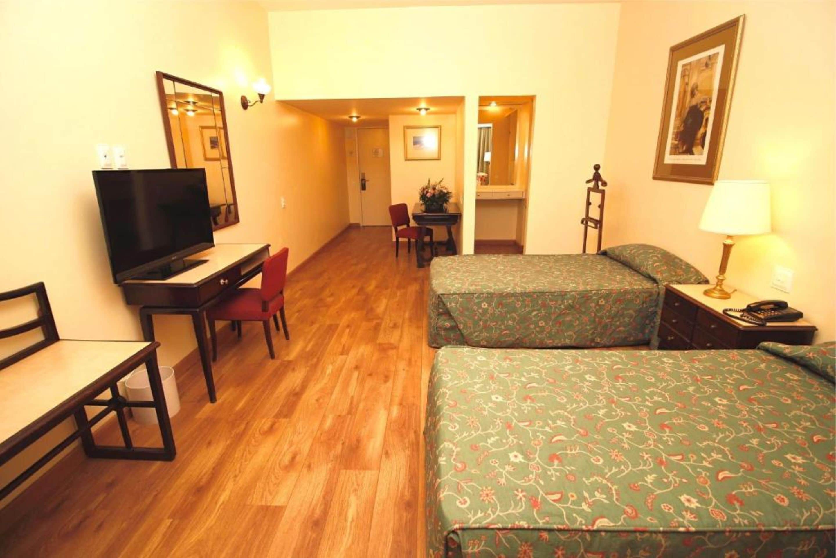 Habitación de hotel con piso de madera, un escritorio con televisor, dos camas con ropa de cama floral, una silla, una mesita de noche con lámpara y un cuadro en la pared.