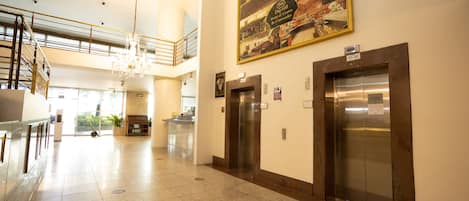 A hotel lobby with a high ceiling, a chandelier, two elevators, a staircase, and a large framed picture on the wall.