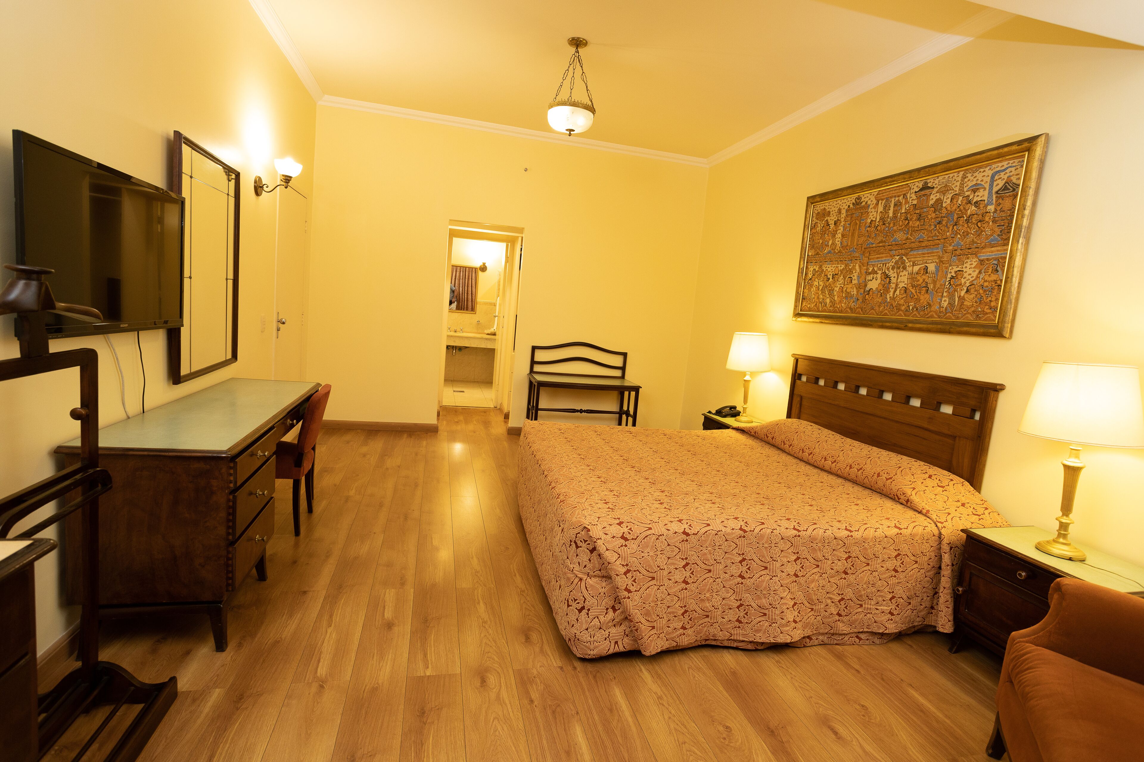 A hotel room with a wooden floor, a bed with a floral bedspread, a desk with a chair, a large mirror, a nightstand with a lamp, and a painting on the wall.